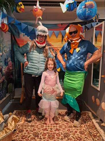 Trois personnes vêtues de costumes colorés sur le thème de la mer posent joyeusement dans une pièce décorée de poissons suspendus, de banderoles et d'un tapis à motifs. L'atmosphère est festive et enjouée.