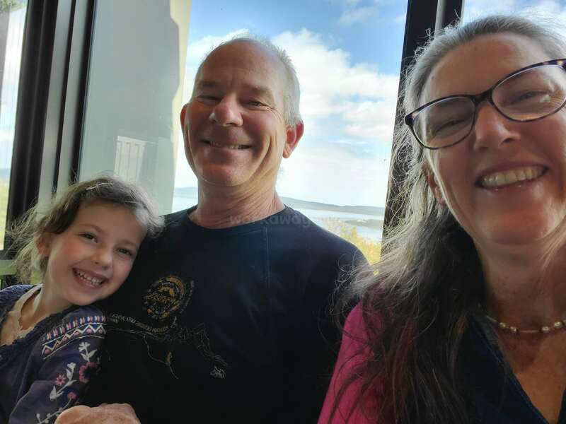 A imagem mostra uma família de três pessoas posando para uma selfie, com uma vista panorâmica do oceano e do céu visível por uma janela atrás deles.