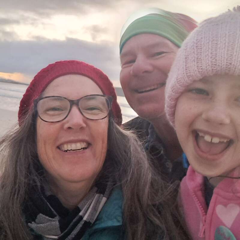 Trois personnes, probablement une famille, emmitouflées dans des bonnets et des vestes d'hiver, profitent d'un moment de joie sur la plage au coucher du soleil, en souriant et en riant joyeusement.