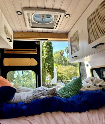 O interior aconchegante da van tem uma cama confortável com almofadas coloridas, armários brancos, um ventilador de teto e uma vista incrível das árvores e do jardim ensolarado lá fora.