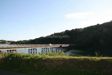 L'image représente un pont enjambant une étendue d'eau, avec une rive herbeuse au premier plan et une colline à l'arrière-plan.