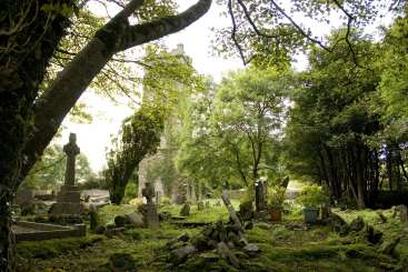 L'image représente un cimetière serein et envahi par la végétation, avec une église en pierre à l'arrière-plan, entourée de verdure luxuriante et de pierres tombales couvertes de mousse.