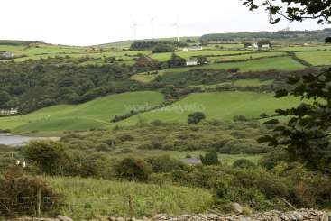 L'image représente un paysage serein de collines ondulantes, de champs verts et d'arbres, avec un plan d'eau au loin, sur fond de ciel couvert.