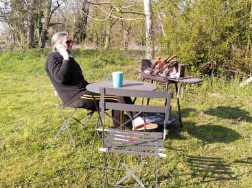Une personne est assise à une table en plein air, au téléphone, avec des lunettes de soleil. Une tasse bleue est posée sur la table, et un barbecue se trouve à proximité.