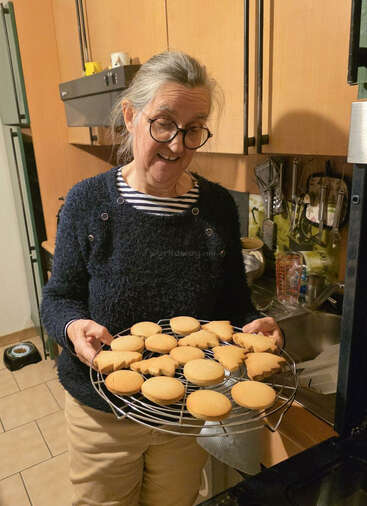 Une femme âgée portant des lunettes tient fièrement une grille de refroidissement sur laquelle sont disposés des biscuits tout juste sortis du four, dont certains ont la forme d'arbres, dans une cuisine chaleureuse dotée d'armoires en bois.