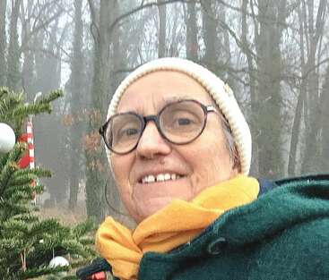Une femme d'un certain âge, portant des lunettes, un bonnet en tricot blanc et une écharpe jaune, sourit chaleureusement. Elle se tient à l'extérieur, dans une forêt brumeuse, près d'un sapin décoré.