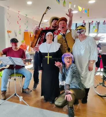 Six adultes vêtus de costumes colorés posent joyeusement à l'intérieur. Des décorations sont suspendues au plafond. L'un d'entre eux joue de la guitare, une autre porte un habit de religieuse, et tout le monde a l'air de faire la fête.