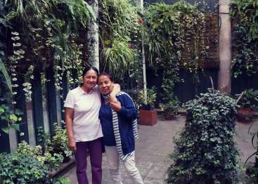 The image depicts two women standing in a lush garden, surrounded by a variety of plants and foliage, with a serene and natural atmosphere.