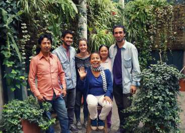 The image depicts a group of six individuals posing for a photo in a lush, verdant setting, surrounded by plants and foliage.