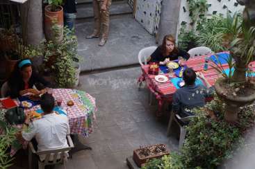 The image depicts a courtyard with four individuals seated at two tables, surrounded by plants and a stone floor, with a subtle watermark in the center.