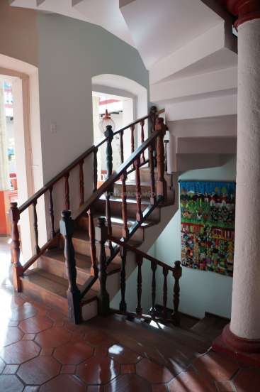 The image depicts a staircase with a wooden railing, situated in a room with white walls and a vibrant tapestry on the wall.