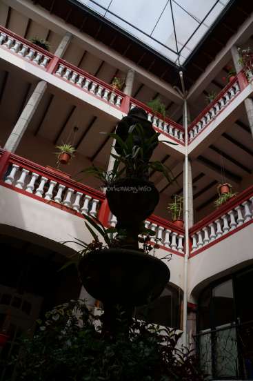 The image depicts a building with a glass roof, featuring a central plant and multiple levels with red and white railings, and a large window on the right.