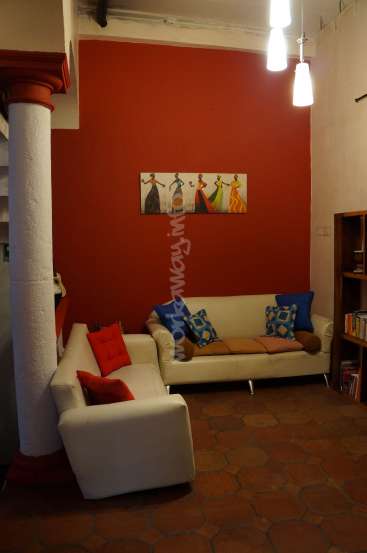 The image depicts a cozy living room with a white couch, red and blue pillows, a red wall, and a white column, featuring a painting of four women dancing.