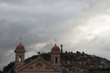 The image depicts a church with two towers and a statue on a hill, set against a cloudy sky. The church is orange and white, with a cross on top of each tower.