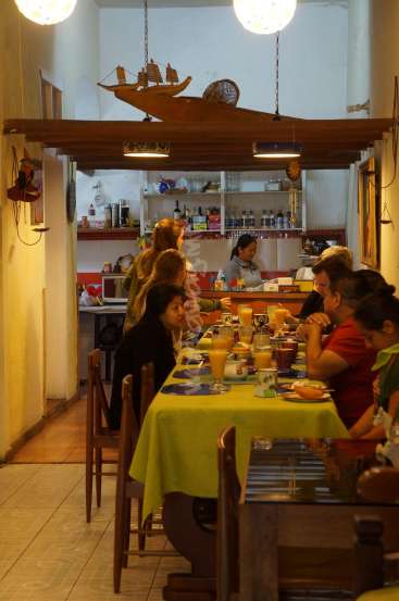The image depicts a restaurant with a long table and chairs, featuring a yellow tablecloth and a bar in the background, with a ship hanging from the ceiling.