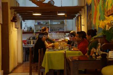The image depicts a restaurant with a group of people seated at a table, featuring a yellow tablecloth and a vibrant mural on the wall.