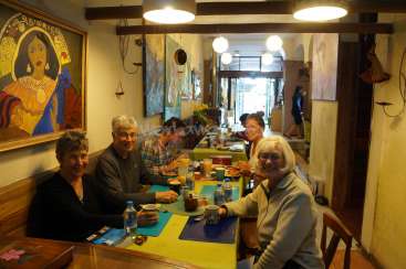 The image depicts a group of people seated at a long table in a restaurant, surrounded by vibrant artwork and warm lighting. The atmosphere appears lively and convivial.
