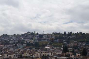 The image depicts a densely populated hillside town, characterized by numerous buildings and trees, set against a cloudy sky.