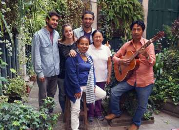 The image depicts a group of six individuals posing for a photo in a lush garden setting, with one person playing a guitar.