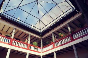 The image depicts a building with a glass roof and a balcony, featuring white pillars and red railings, adorned with potted plants.