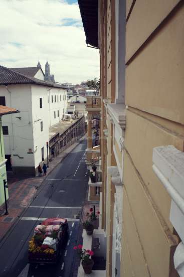 The image depicts a narrow street in an urban area, with a yellow building on the right and a red truck carrying flowers on the left.