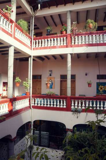 The image depicts a multi-story building with a balcony, featuring white and red railings, and potted plants hanging from the railings.
