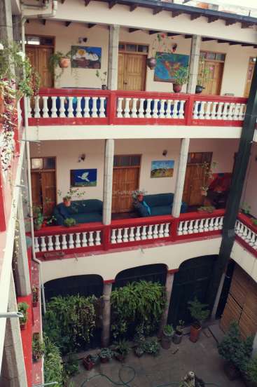 The image depicts a three-story building with a balcony on each floor, featuring a red railing and white balusters, surrounded by potted plants and artwork.