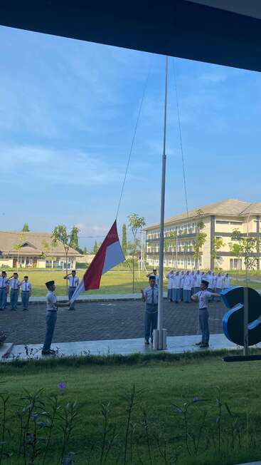 Das Bild zeigt eine Gruppe von Menschen, die die indonesische Flagge an einem Mast hissen, mit einem großen Gebäude im Hintergrund und einem blauen Himmel darüber.