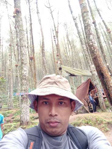 Ein Mann mit Eimerhut und Rucksack macht ein Selfie in einem Wald mit hohen, dünnen Bäumen, einer Holzhütte und ein paar Menschen im Hintergrund.