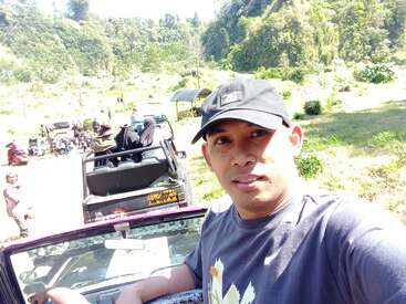 Ein Mann mit einer schwarzen Mütze und einem T-Shirt macht ein Selfie im Freien. Hinter ihm sind mehrere Menschen und Jeeps in einer malerischen, grünen Landschaft zu sehen.