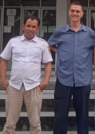 Dos hombres están uno al lado del otro, sonriendo frente a la entrada de un edificio. Uno lleva un uniforme blanco y el otro una camisa azul, y ambos transmiten un ambiente de cordialidad y camaradería.
