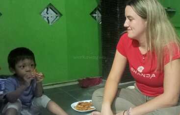 Una joven y un niño están sentados en el suelo dentro de una casa, compartiendo la comida. La mujer sonríe con cariño mientras el niño come, creando un ambiente acogedor.