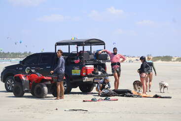 A group of people and dogs gather on a sandy beach beside a black truck and red ATV, preparing kitesurfing gear under a clear, sunny sky.