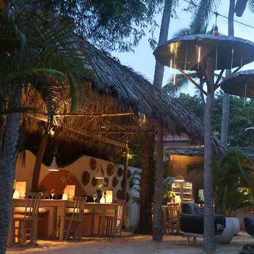 A cozy outdoor restaurant with thatched roofs, wooden furniture, warm lighting, and tropical plants. Evening ambiance enhanced by hanging lamps and a relaxed, inviting atmosphere.