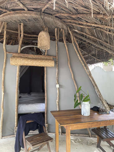 This rustic hut features a wooden table with a leafy plant in a jar, simple chairs, a bed inside, natural wood supports, and a thatched roof.