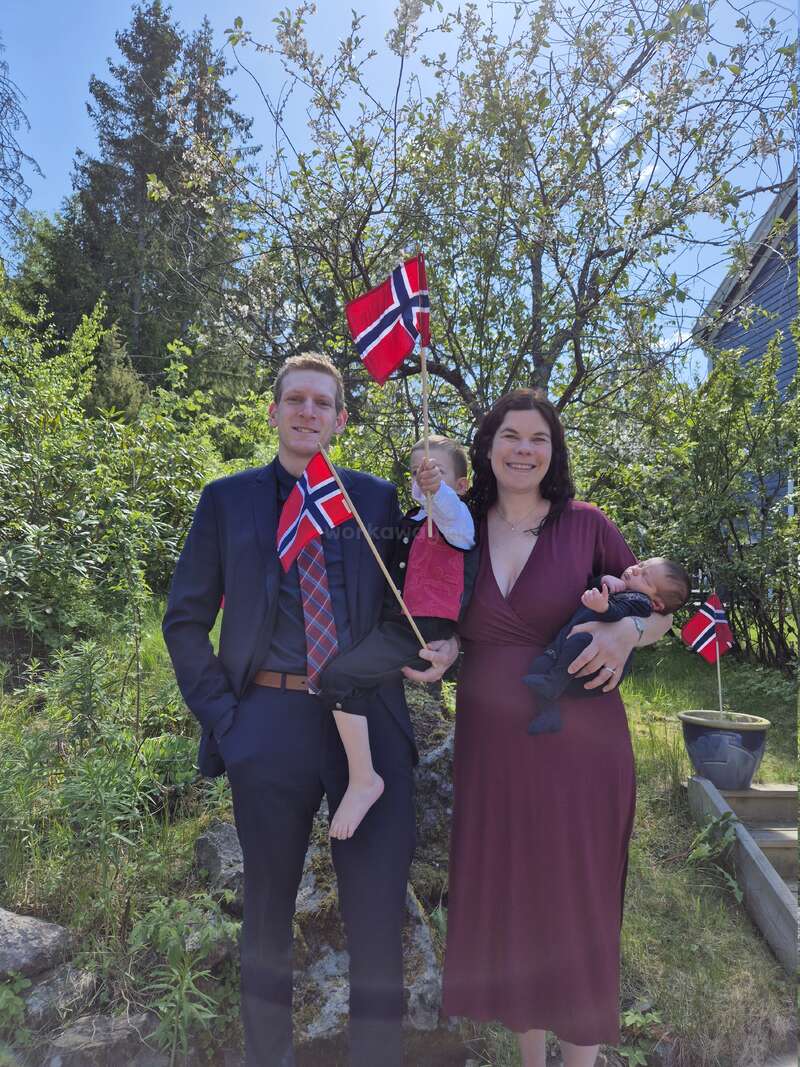 Uma família sorridente de quatro pessoas está ao ar livre, sob o sol, segurando bandeiras da Noruega. Cercados por vegetação, eles parecem felizes e bem vestidos, comemorando juntos uma ocasião especial.