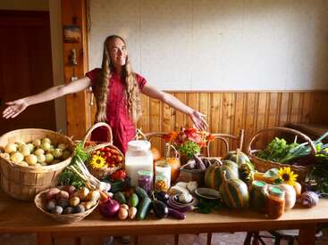 Une femme souriante se tient derrière une table débordant de légumes colorés du potager, de fruits, de bocaux, de fleurs et de paniers de produits frais, présentant fièrement une récolte abondante à l'intérieur.