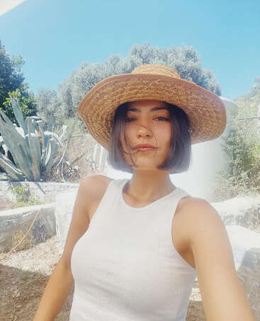 A woman with a bob haircut wears a wide-brimmed straw hat and white tank top, taking a selfie outdoors on a sunny day with green trees behind.