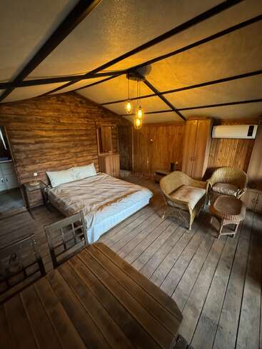 This cozy wooden cabin bedroom features a double bed, wicker chairs, a small table, wardrobe, rustic wooden floors, warm lighting, and an air conditioner on the wall.