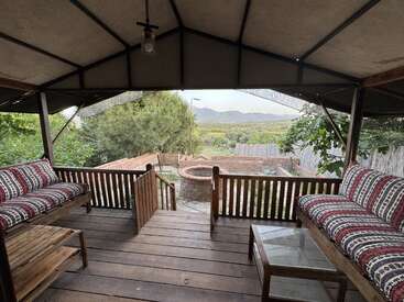 A cozy wooden gazebo features patterned cushioned benches, a glass coffee table, and overlooks a green garden with a stone well and distant mountains visible.