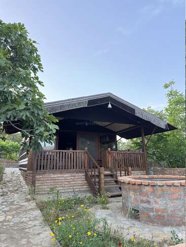 A rustic wooden cabin with a shaded porch sits amidst greenery and flowers. A stone path and circular brick structure enhance the cozy, serene countryside setting.