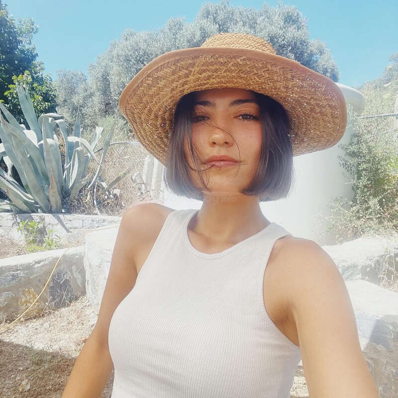 A woman with a bob haircut wears a wide-brimmed straw hat and white tank top, taking a selfie outdoors on a sunny day with green trees behind.