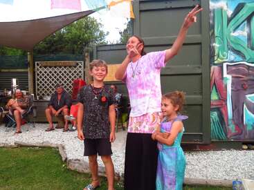 L'image représente une femme et deux enfants debout à l'extérieur, la femme faisant le signe de la paix, vêtue d'une chemise et d'une jupe longue à motif de cravate.