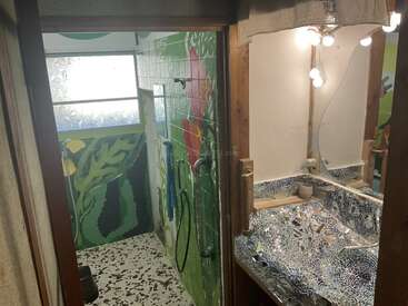 Esse banheiro eclético tem murais vibrantes inspirados na natureza, piso estilo terrazzo, pia com azulejos em mosaico, lâmpadas expostas e janelas foscas pra uma luz natural suave e difusa.