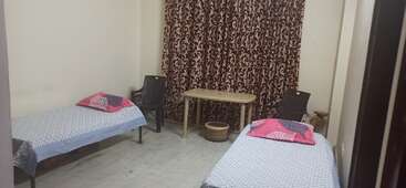 The image depicts a room with two beds, a table, and a chair, featuring a white wall and a window, likely a bedroom or living space. The room is well-lit and organized.