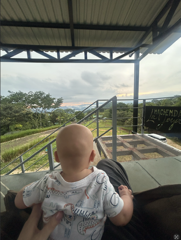 Un bebé sentado en el regazo de alguien bajo una terraza cubierta, contemplando un paisaje verde y tranquilo y una puesta de sol lejana, creando un momento sereno y acogedor juntos.