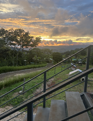 Una escalera metálica domina un exuberante paisaje verde bajo un vibrante y dramático cielo al atardecer. Las nubes brillan en naranja y amarillo sobre montañas lejanas y un apacible paisaje rural.