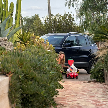 Una mujer de pelo largo y rubio está agachada junto a un todoterreno negro, jugando con un niño en un coche rojo de juguete en una entrada rodeada de plantas.