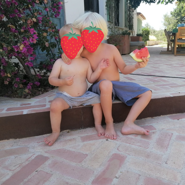 Dos niños pequeños, uno de ellos con una sandía en la mano, se sientan juntos en un escalón de ladrillo frente a una casa, con sus rostros difuminados para mayor intimidad.