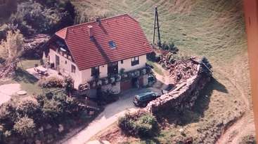 The image depicts a large house with a gable roof, featuring a spacious yard and a driveway, surrounded by trees, with a serene and peaceful atmosphere. The house is situated in a rural area.